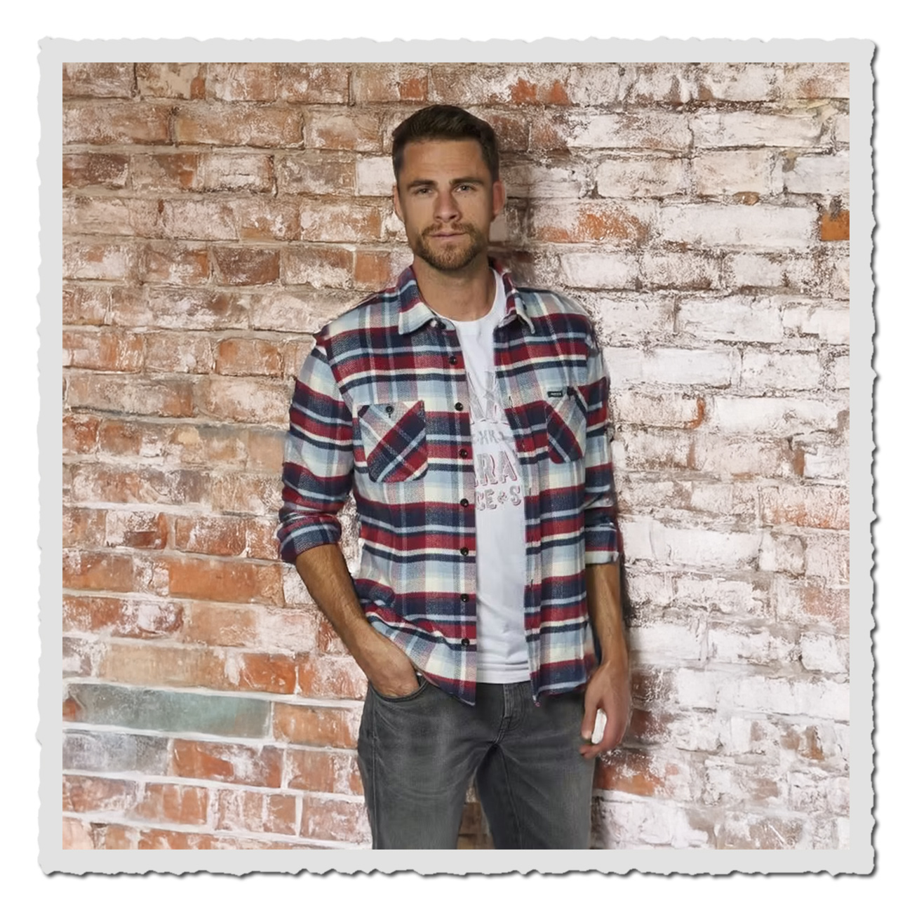 Mann trägt The Rokker Company Flanellhemd Orlando in Rot-Blau-Weiß-Karo mit T-Shirt und Jeans vor einer Klinkerwand